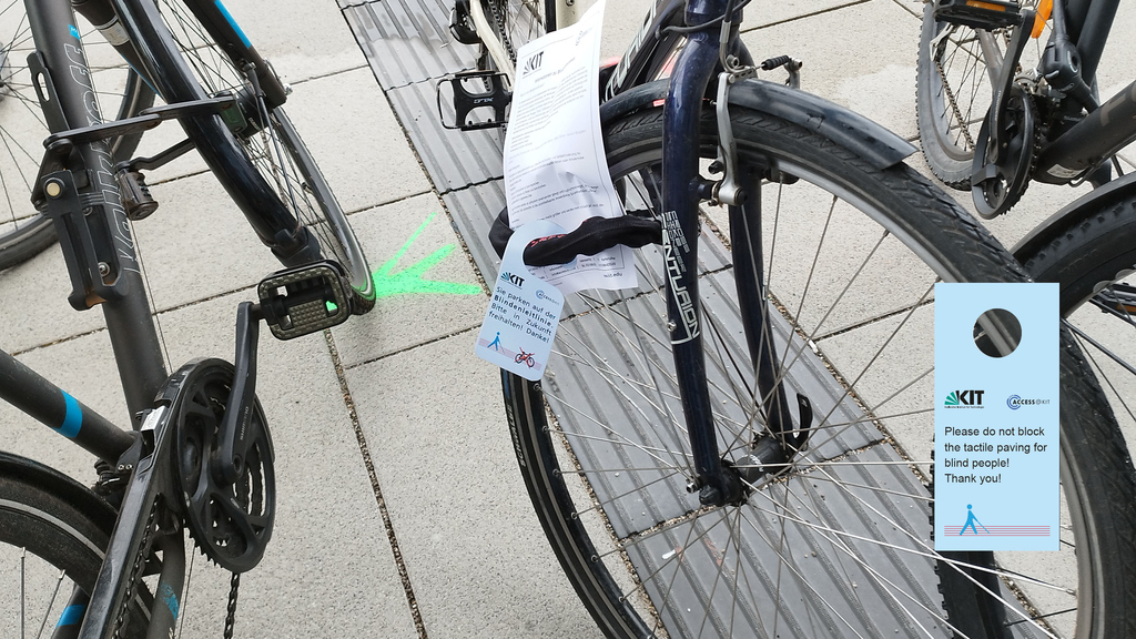 Das Foto zeigt ein Fahrrad, das auf dem Blindenleitsystem abgestellt ist. Eine Markierung auf dem Boden weist wie auch ein Informationsanhänger am Rad darauf hin, dass das Blindenleitsystem freigehalten werden soll.